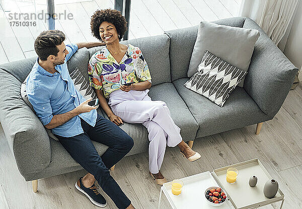 Glückliches multiethnisches Paar hält Smartphones in der Hand  während es auf dem Sofa im Penthouse sitzt