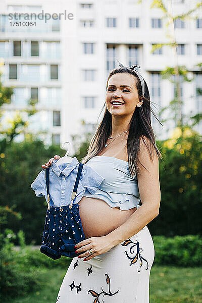 Lächelnde schwangere Frau hält Babykleidung in der Hand  während sie im Park steht