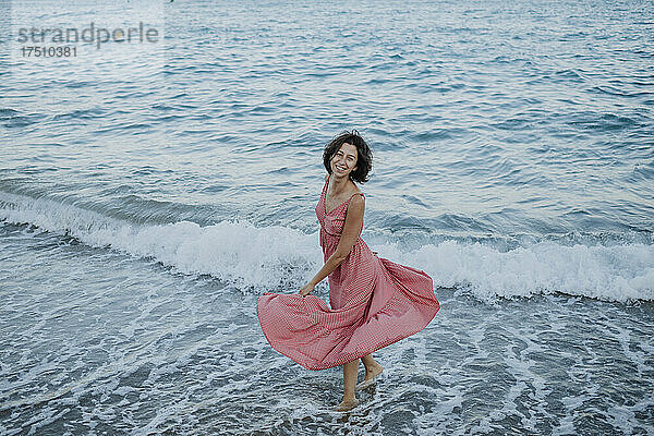 Fröhliche Frau in Kleid steht bei Sonnenuntergang im Meer