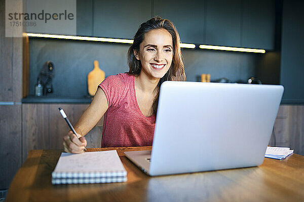 Lächelnde Unternehmerin schaut auf den Laptop  während sie in Notizblock auf dem Schreibtisch schreibt