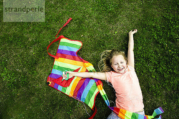 Fröhliches Mädchen hält Drachen in der Hand  während es im Park auf Gras liegt