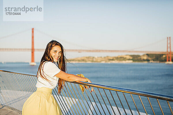 Lächelnde schöne Frau mit langen Haaren  die bei Sonnenuntergang am Geländer über dem Fluss in der Stadt steht