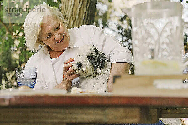 Lächelnde Seniorin spielt mit Hund  während sie am Tisch im Garten sitzt