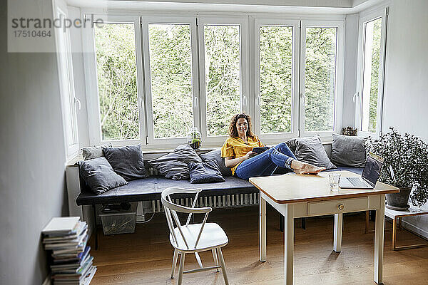 Frau mit digitalem Tablet entspannt sich zu Hause auf dem Sofa vor dem Fenster