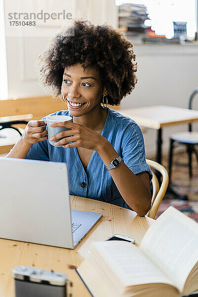 Nachdenkliche Frau hält Kaffeetasse in der Hand  während sie mit Laptop im Café sitzt