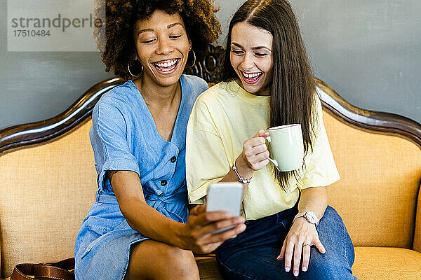 Fröhliche Frauen machen Selfies per Smartphone  während sie auf dem Sofa in einem modernen Café sitzen