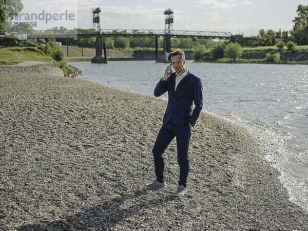 Geschäftsmann spricht über Smartphone  während er an sonnigen Tagen am Flussufer steht
