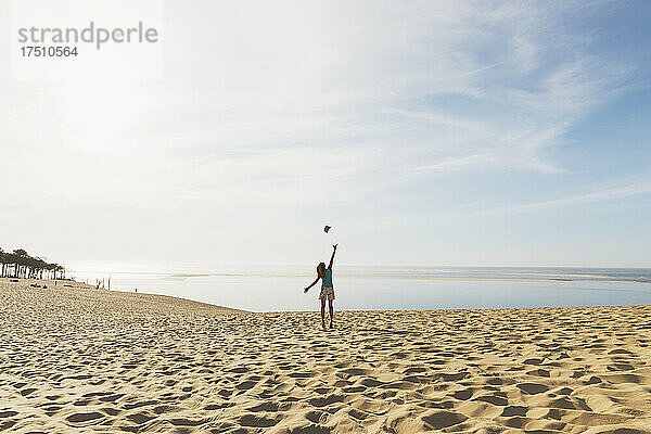 Glückliche Frau wirft Hut in die Luft  während sie am Strand gegen den Himmel steht  Düne von Pilat  Nouvelle-Aquitaine  Frankreich
