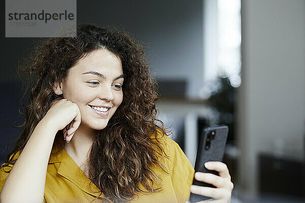 Lächelnde schöne Frau  die ihr Smartphone benutzt  während sie zu Hause sitzt