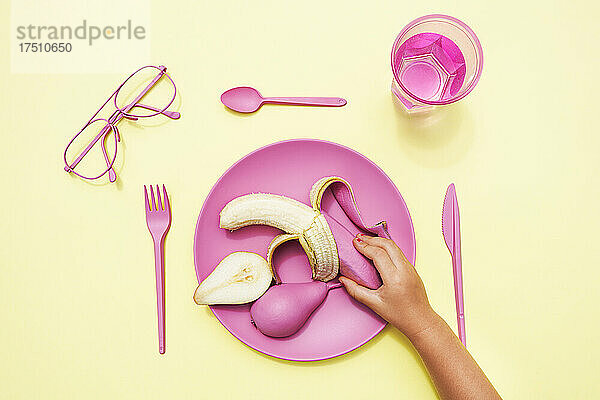 Hand eines kleinen Mädchens  das eine rosafarbene Banane von einem Plastikteller aufnimmt