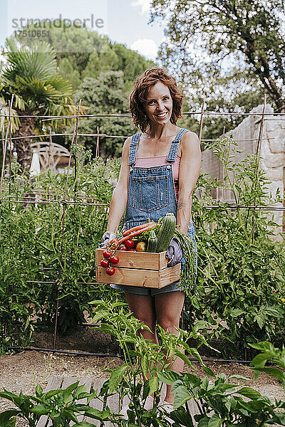 Lächelnde Frau hält verschiedene Gemüsesorten in einer Kiste  während sie im Gemeinschaftsgarten vor Pflanzen steht