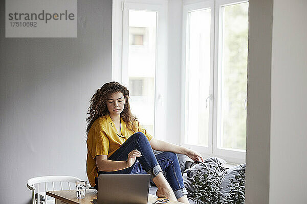 Junge Frau sitzt am Tisch und benutzt Laptop für Online-Hausaufgaben zu Hause