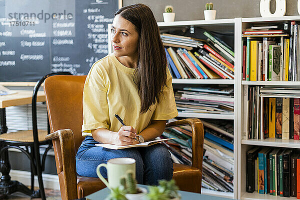Nachdenkliche Frau schreibt in ein Buch  während sie auf einem Stuhl im Café sitzt