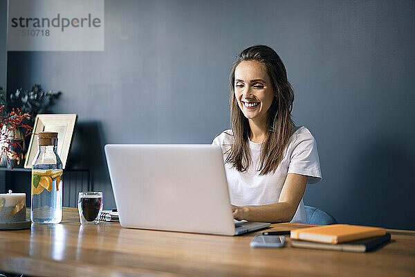 Lächelnde junge Frau benutzt Laptop auf Schreibtisch an der Wand im Heimbüro