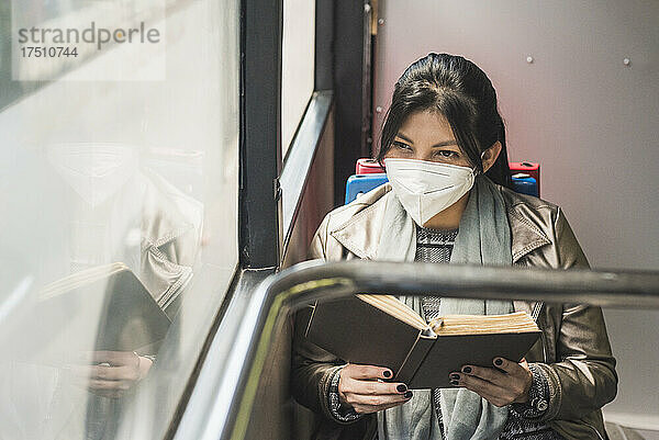 Frau trägt Schutzmaske und schaut durch das Fenster  während sie im Bus ein Buch liest