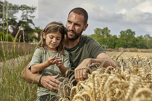 Vater und Tochter sitzen neben Feldfrüchten im Weizenfeld vor dem Himmel