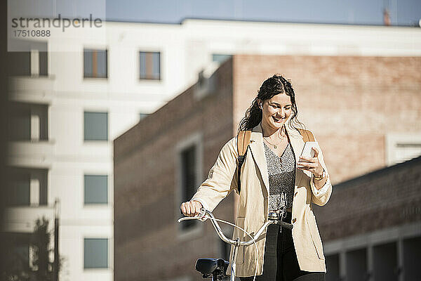Lächelnde Pendlerin nutzt ihr Mobiltelefon  während sie mit dem Fahrrad gegen Gebäude in der Stadt läuft