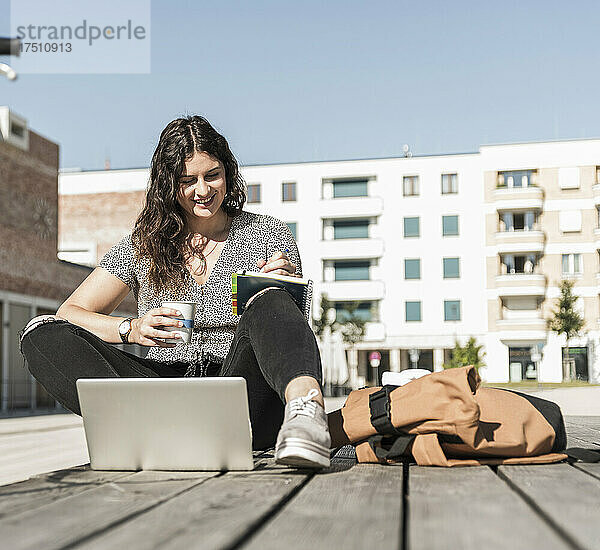 Lächelnde Frau hält ein Getränk in der Hand und schreibt in ein Buch  während sie an einem sonnigen Tag auf der Promenade sitzt