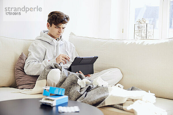 Kranker junger Mann nutzt digitales Tablet  während er sich auf dem Sofa im Wohnzimmer entspannt