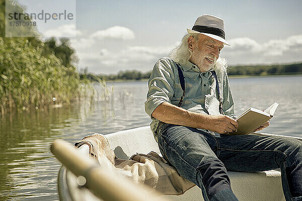 Porträt eines zufriedenen älteren Mannes  der im Ruderboot auf einem See sitzt und ein Buch liest