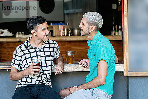 Lächelndes schwules Paar unterhält sich  während es an der Bartheke Bier trinkt