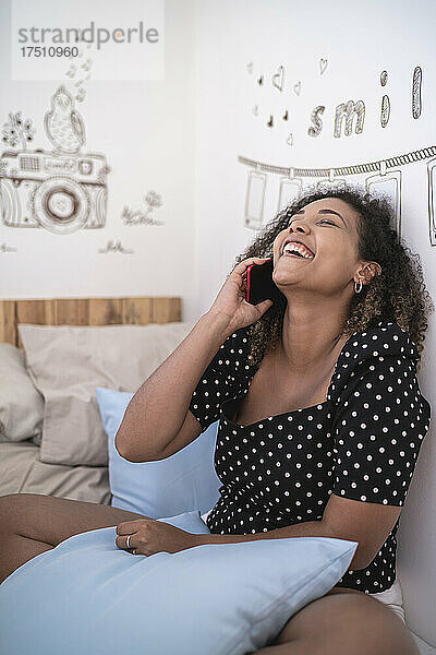 Fröhliche Frau mit lockigem Haar  die zu Hause an der Wand sitzt und über ihr Smartphone spricht