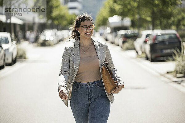 Lächelnde junge Frau mit Tasche  die an einem sonnigen Tag auf der Straße läuft