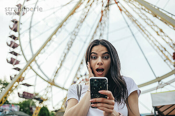 Überraschte junge Frau mit offenem Mund benutzt Smartphone gegen Riesenrad im Vergnügungspark