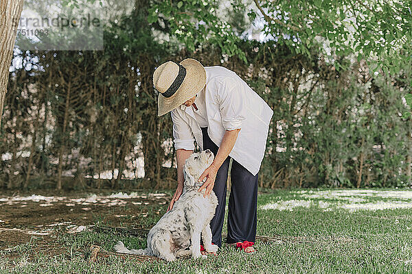 Ältere Frau mit Hut und Hund steht auf Grasfläche im Hof