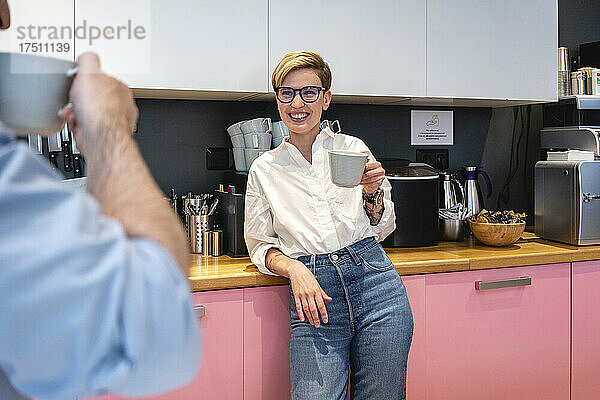 Lächelnde schöne Geschäftsfrau hält Kaffeetasse in der Hand  während sie ihren männlichen Kollegen während der Pause in der Bürokantine ansieht