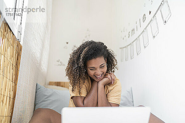 Fröhliche Frau mit lockigem Haar führt Videokonferenzen über Laptop  während sie zu Hause an der Wand sitzt