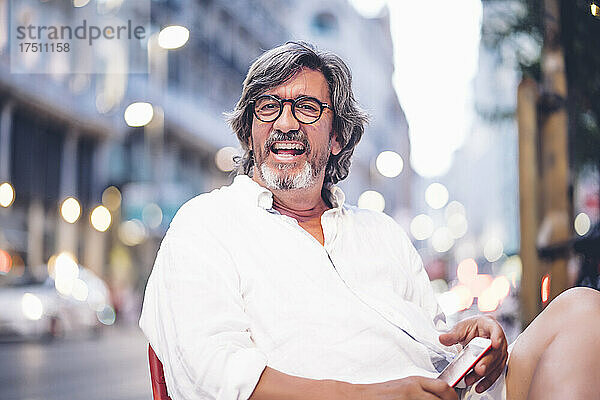 Porträt eines fröhlichen älteren Mannes  der mit seinem Smartphone im Straßencafé in Madrid sitzt