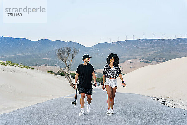 Lächelndes multiethnisches Paar  das auf der Straße inmitten von Sanddünen vor klarem Himmel spaziert