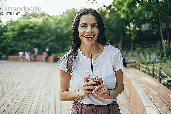Fröhliche junge Frau hält einen Erfrischungsgetränkebecher in der Hand  während sie im Park steht