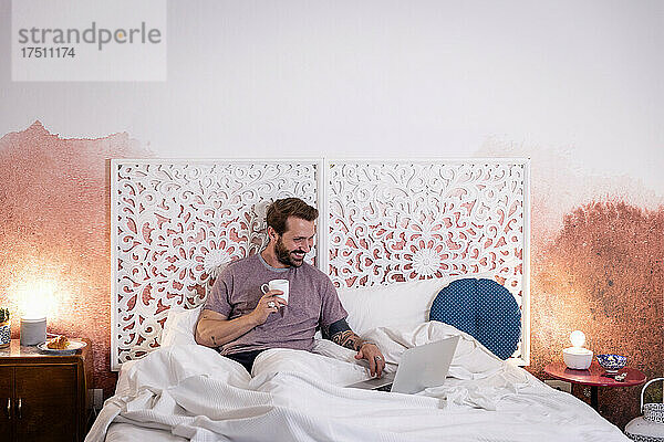 Lächelnder erwachsener Mann hält Kaffee mit Laptop in der Hand und entspannt sich zu Hause im Bett