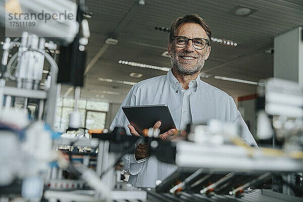 Lächelnder männlicher Wissenschaftler hält digitales Tablet in der Hand  während er neben Maschinen im Labor steht