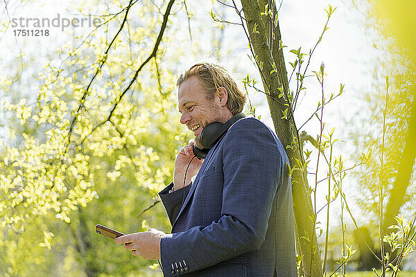 Lächelnder Geschäftsmann benutzt Smartphone  während er an einem sonnigen Tag im Park steht