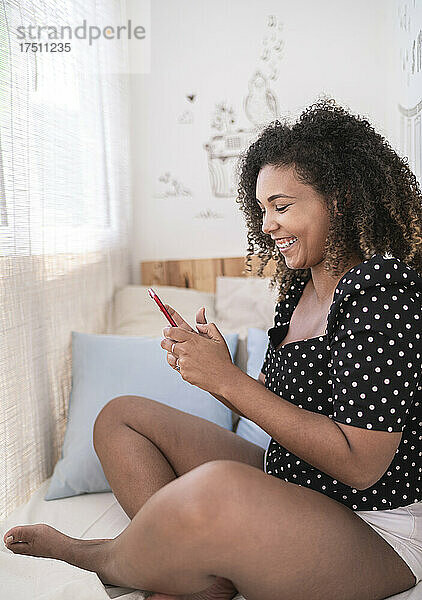 Lächelnde Frau mit lockigem Haar benutzt Smartphone  während sie zu Hause im Bett sitzt