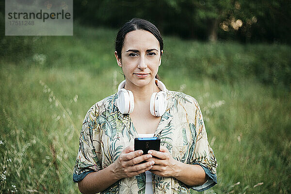 Frau steht mit Smartphone und Kopfhörern im öffentlichen Park