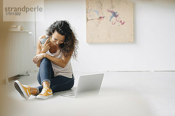 Lächelnde Frau hält Kaffeetasse in der Hand und sitzt zu Hause mit Laptop auf dem Boden