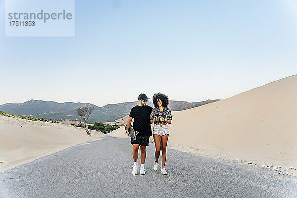 Junges Paar läuft auf der Straße an einer Sanddüne vor dem klaren blauen Himmel