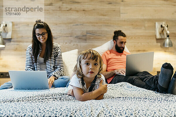 Nettes Mädchen liegt auf dem Bett  während die Mutter am Laptop arbeitet und der Vater zu Hause im Schlafzimmer eingeschlafen ist