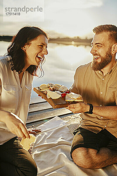 Glückliches Paar beim Picknick am Steg an einem See bei Sonnenuntergang und Käse essend