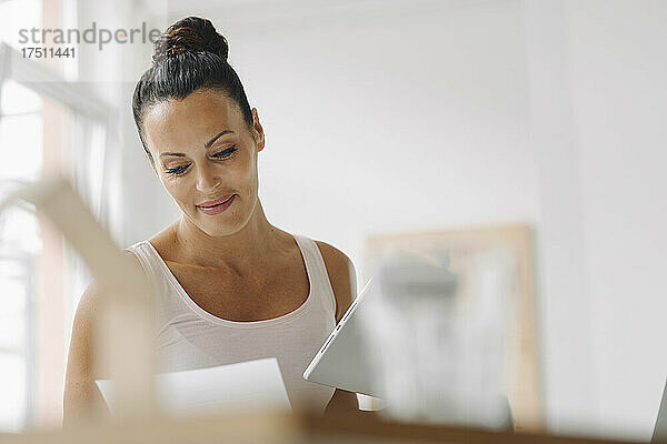 Lächelnde Frau hält ein digitales Tablet in der Hand und liest ein Dokument  während sie im Heimbüro sitzt
