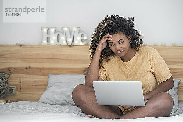 Junge Frau mit lockigem Haar benutzt Laptop  während sie im Bett sitzt