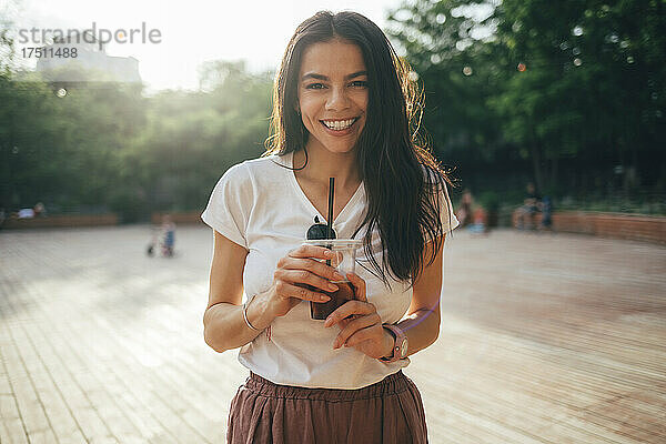 Fröhliche schöne Frau mit langen Haaren hält alkoholfreies Getränk in der Hand  während sie im Park steht