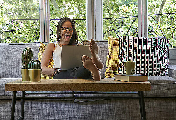 Fröhliche Frau benutzt Laptop  während sie auf dem Sofa im Wohnzimmer sitzt
