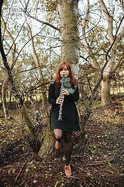 Schöne junge Frau übt Klarinette  während sie sich im Wald auf einen kahlen Baumstamm stützt