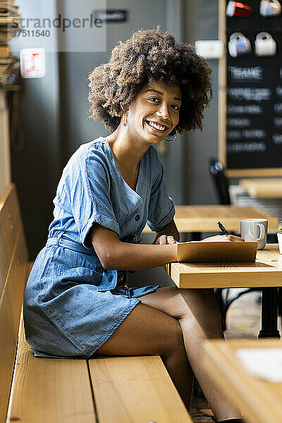 Glückliche junge Frau schreibt am Tisch im Café in ein Buch