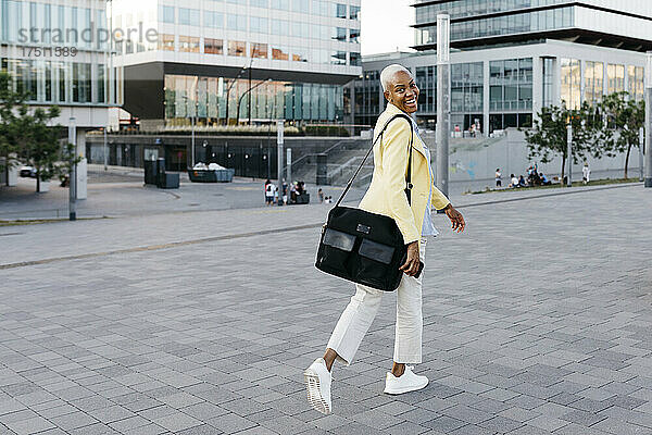 Lächelnde Geschäftsfrau mit Telefon und Tasche spaziert durch die Stadt
